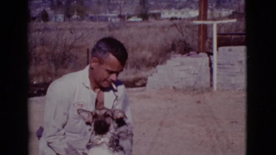The man crouches with the dog facing forward, cigarette still in his mouth, in the 1968 Cottonwood yard scene.