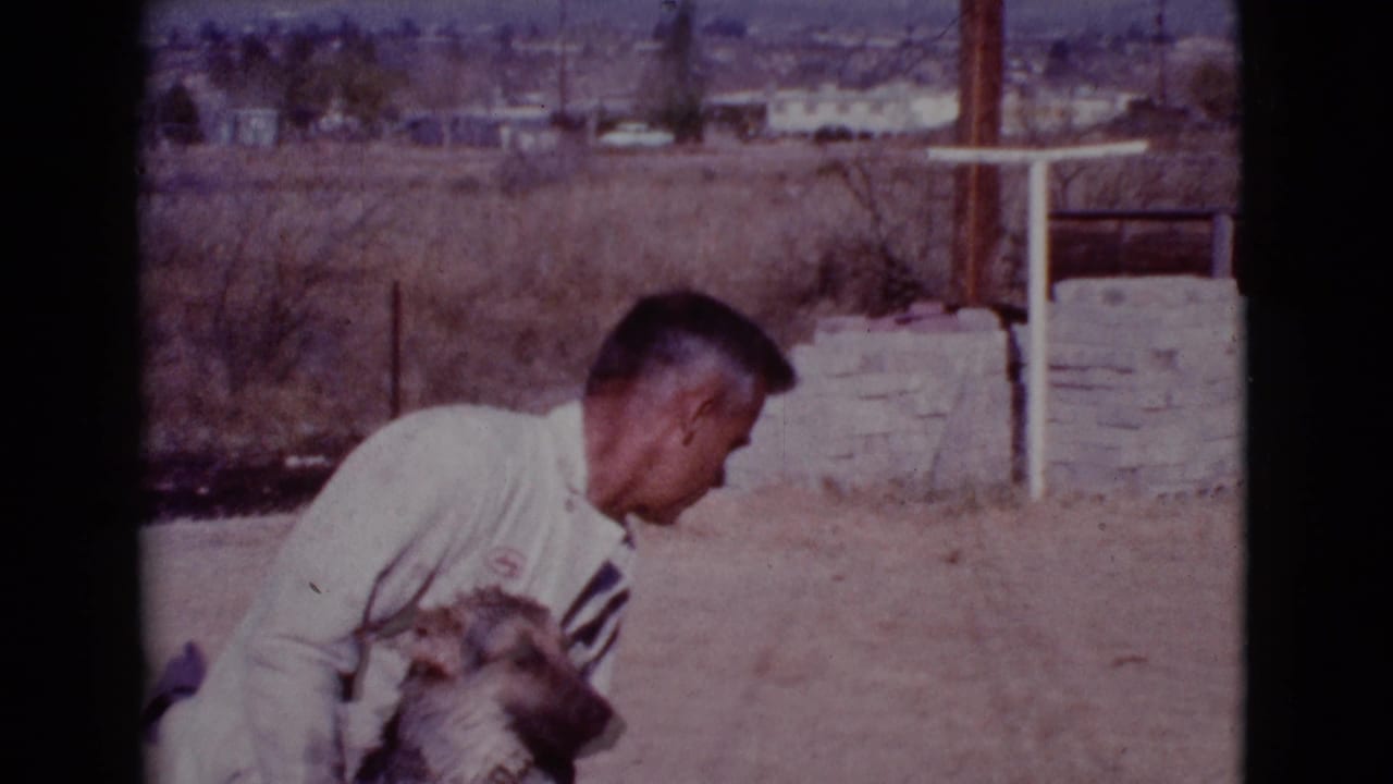 Man crouching beside a dog in a dry yard with a low stone wall behind him.