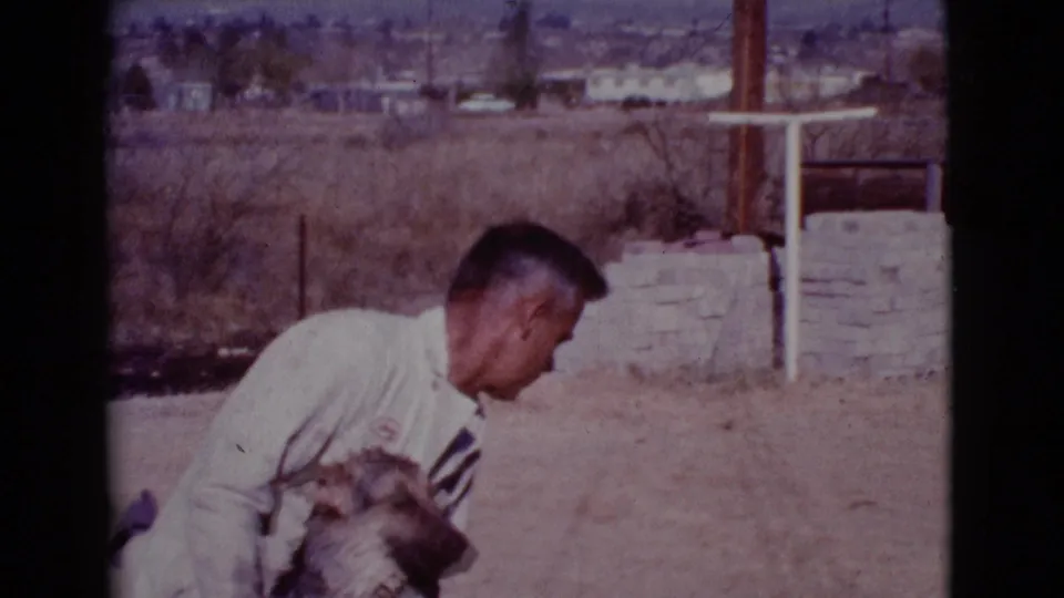 In Cottonwood, Arizona in 1968, a man in a light shirt crouches next to a medium-sized dog in a dry suburban yard.