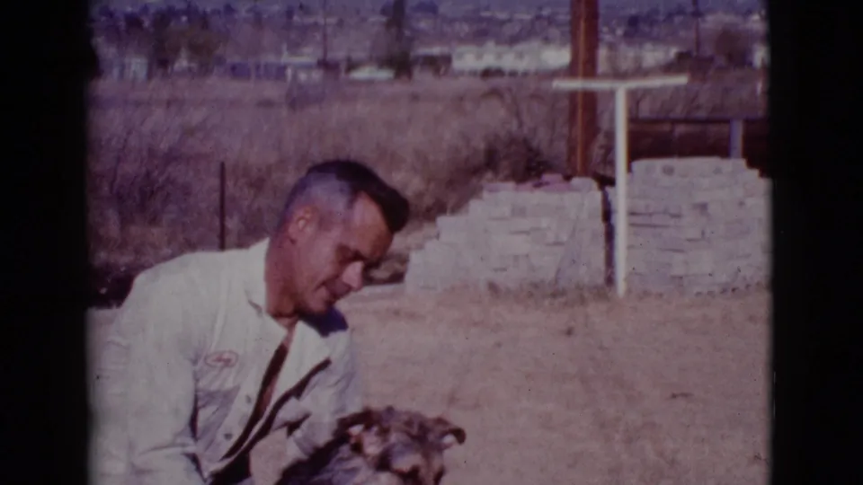 The man bends toward the dog in the 1968 Cottonwood yard, with dry grass, a wall, and distant homes in the background.