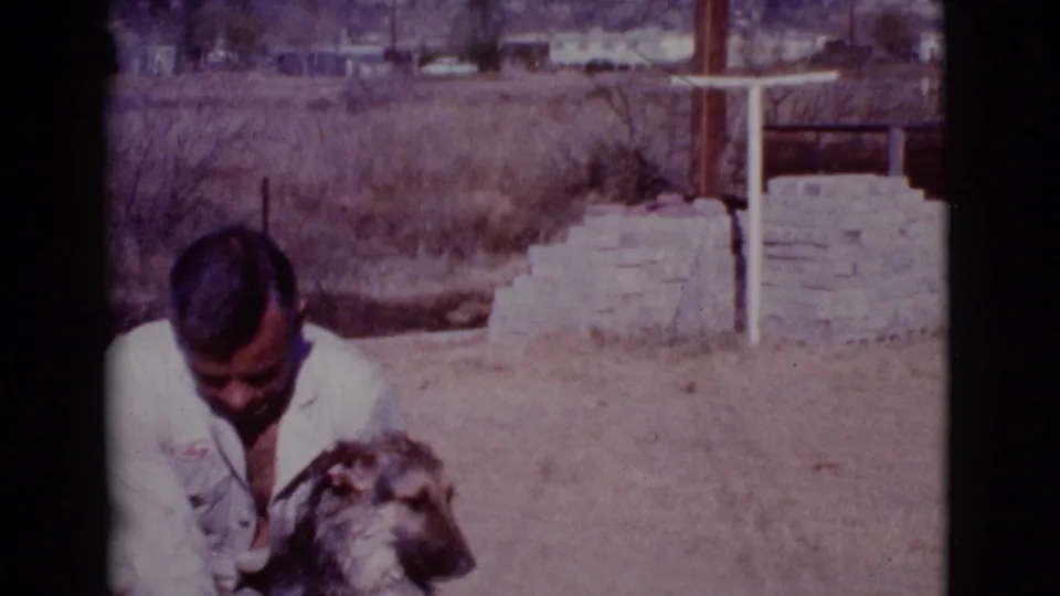 The man and dog remain close together in the Cottonwood, Arizona yard, framed by dry ground, a low wall, and distant houses.