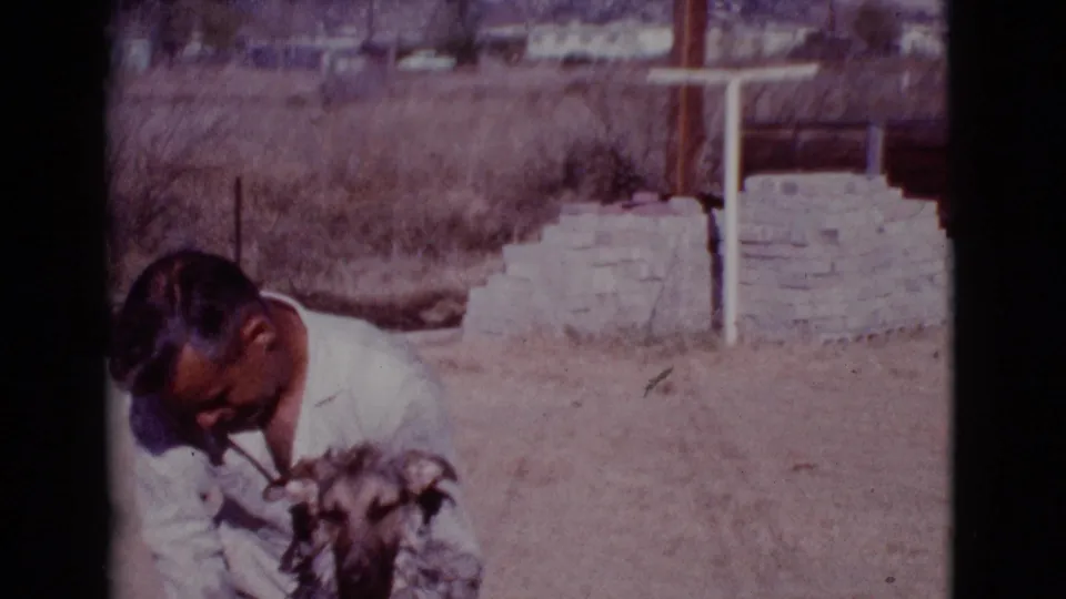 In Cottonwood, Arizona in 1968, a man in a white shirt crouches close to a German Shepherd in a dry outdoor yard.