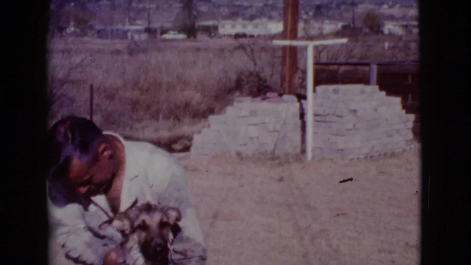 The man bends toward the German Shepherd in a sunlit Cottonwood yard, with dry grass, stacked stone, and distant homes behind them.