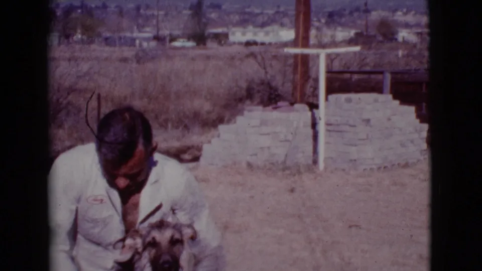 A man in a white shirt looks down toward the German Shepherd during an outdoor washing moment in the 1968 Arizona home-movie footage.