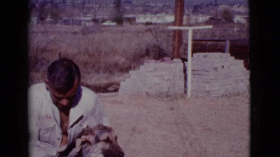 The man remains crouched over the German Shepherd in the Cottonwood yard, framed by dry grass, stonework, and distant houses.