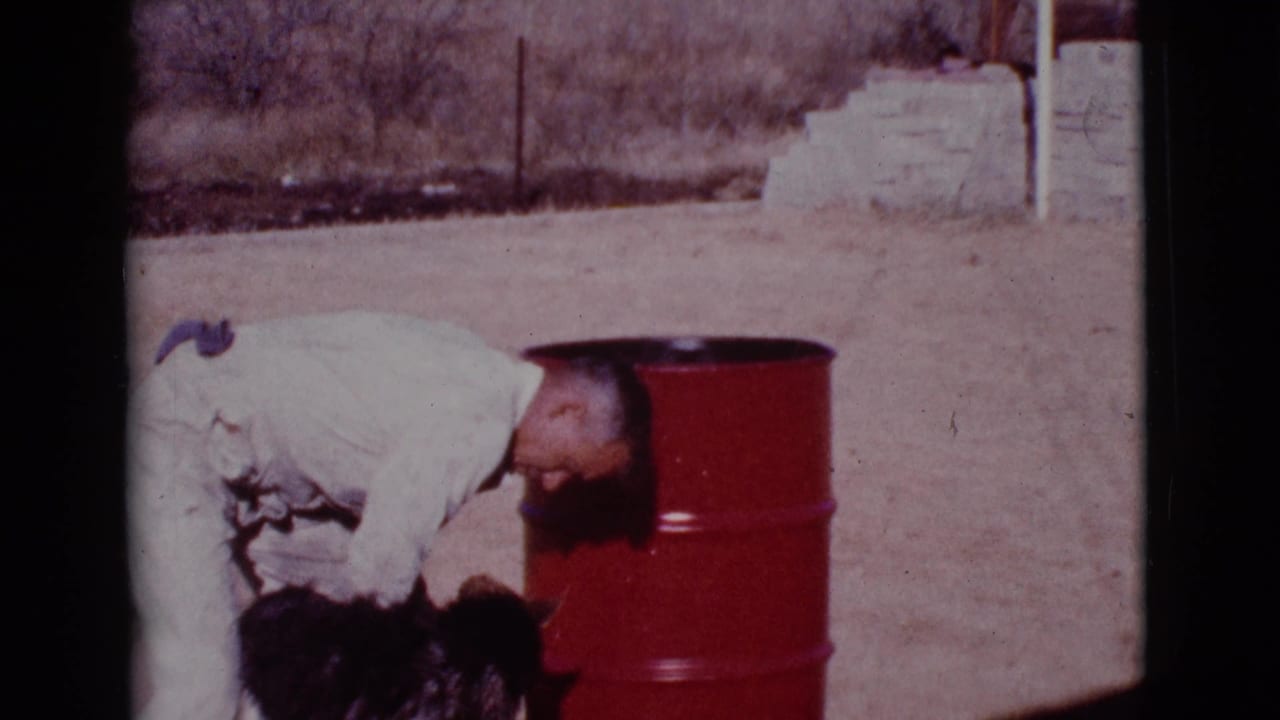 Man bends over a dark dog beside a red barrel in a dusty yard.