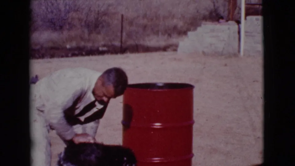 The man stands bent at the waist, looking down at the dark dog beside a red barrel in the Cottonwood yard.