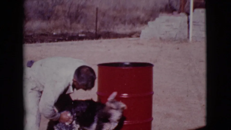 The man leans close over the dark dog, whose head is partly raised, with the red barrel and dry yard behind them.