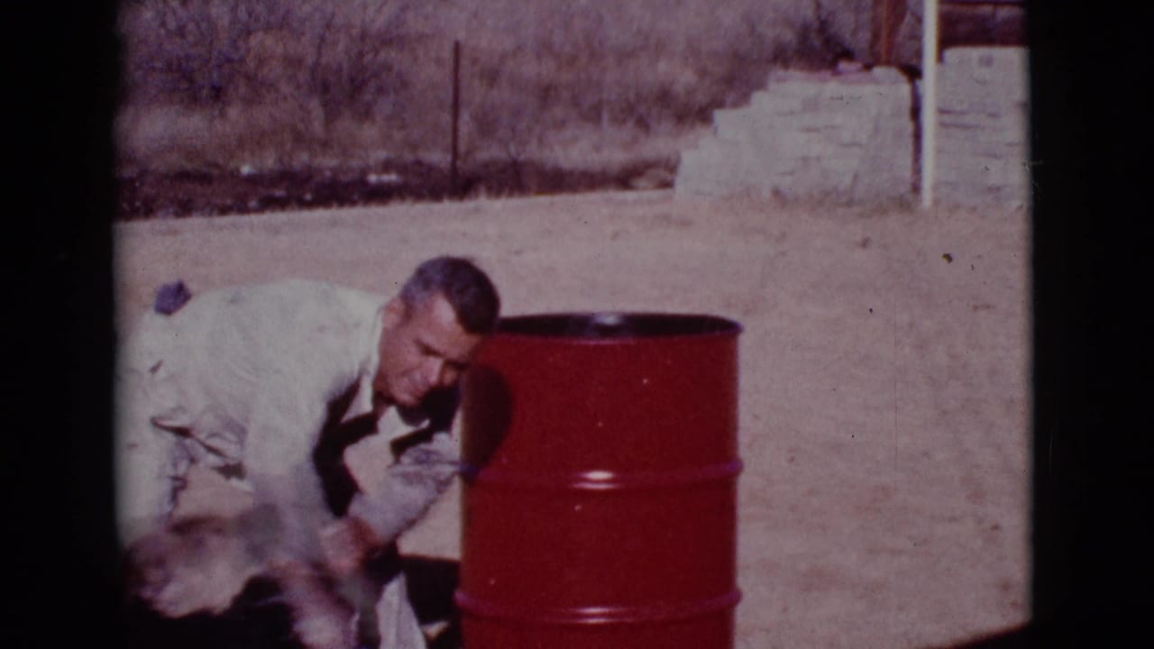 Uniformed man crouches beside a black dog near a red barrel outdoors.