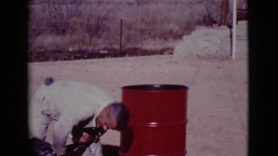 A 1968 home-movie frame shows the uniformed man leaning over the black dog, with a red barrel and dry brush behind them.