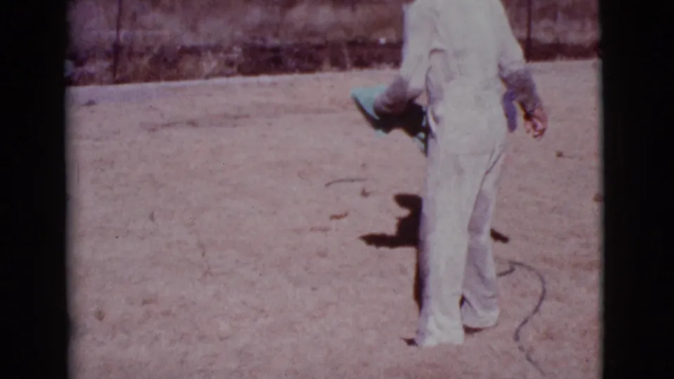 A man in white clothing holds a pale blue cloth in the 1968 Cottonwood yard scene as the play continues off frame.
