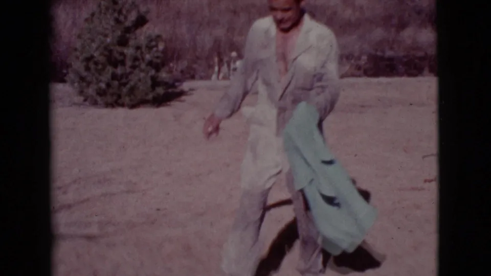 A man in light coveralls carries a pale blue towel through the sunlit Cottonwood yard, with shrubs and sandy ground behind him.