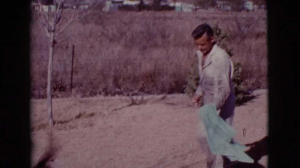 The man carries the green towel through the dusty Cottonwood yard, with sparse vegetation and sharp sunlight visible behind him.