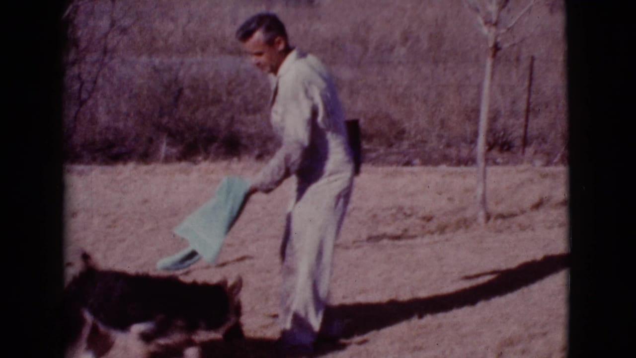 Man in light coveralls holds a green towel while a dark dog walks beside him in a dry yard.