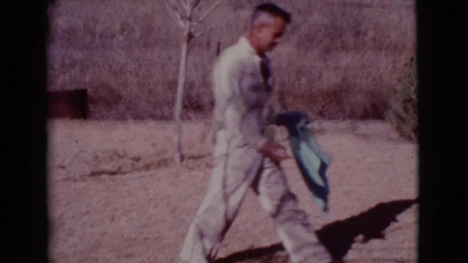 The man continues across the dusty Cottonwood yard holding a green towel, with sparse vegetation and a dog partly visible near the edge of frame.