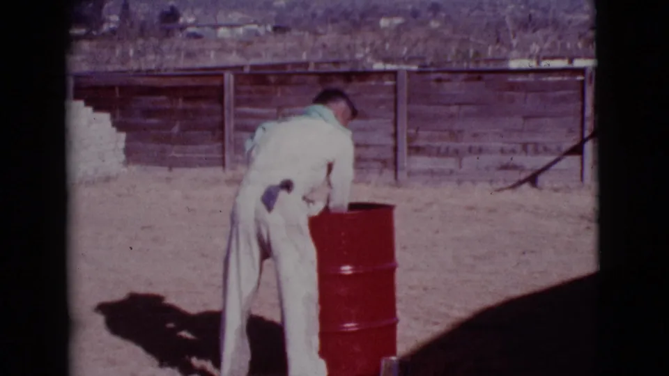 The man bends toward a red metal barrel in the Cottonwood yard, with a weathered wooden fence and dry ground behind him.
