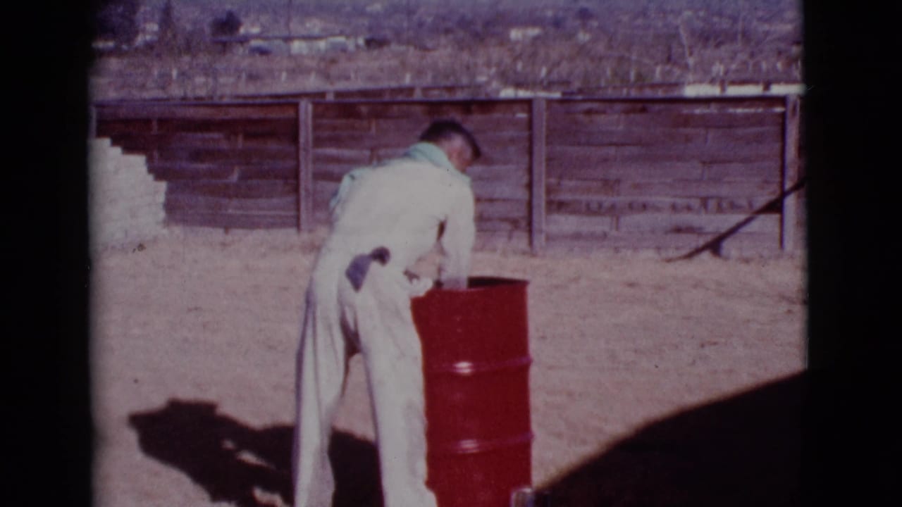 Man in a white jumpsuit bends over a red barrel in a dusty fenced yard.