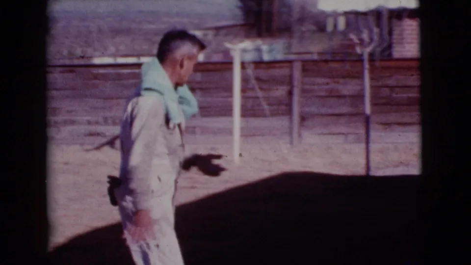 The man walks through the dusty Cottonwood, Arizona yard, with a wooden fence and small trees visible behind him.