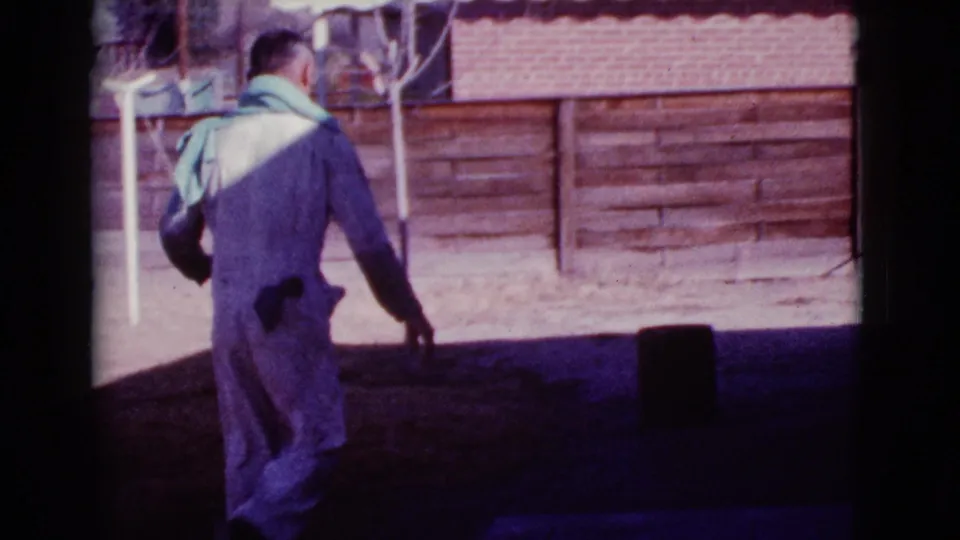 The man turns away in a white jumpsuit as he moves through shadow, with the red barrel and wooden fence behind him.