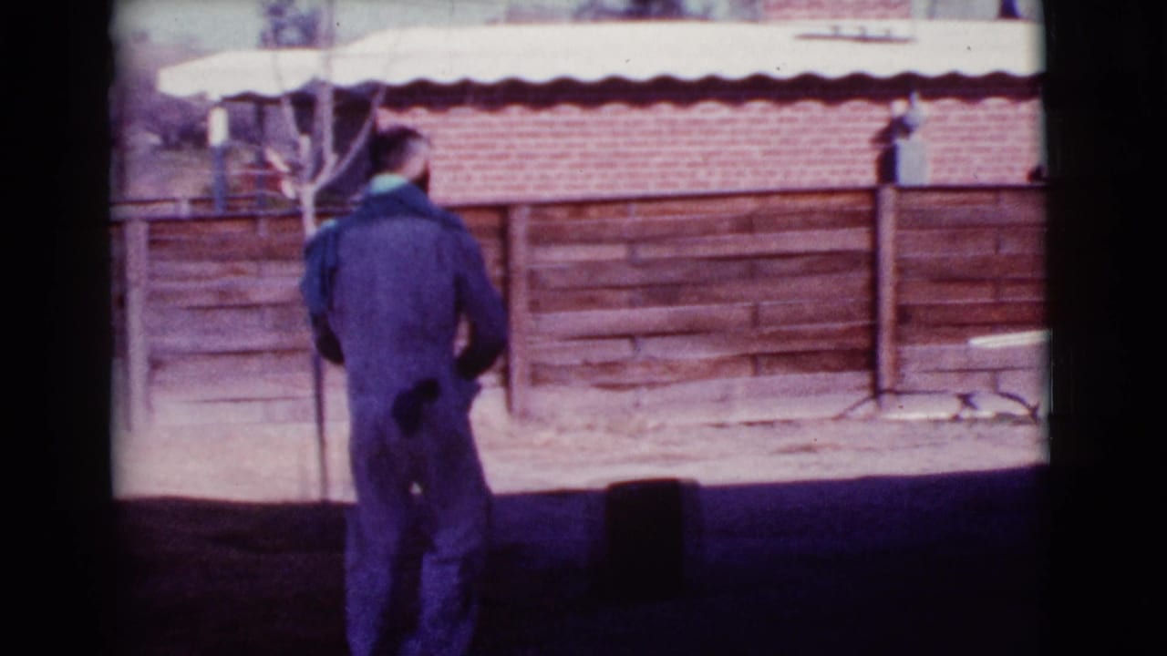 Man in dark or shadowed coveralls stands in a sunny yard near a wood fence.