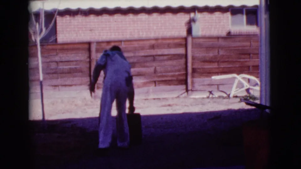 The man bends toward a red barrel in a Cottonwood backyard, with a wooden fence, brick wall, and patio furniture behind him.