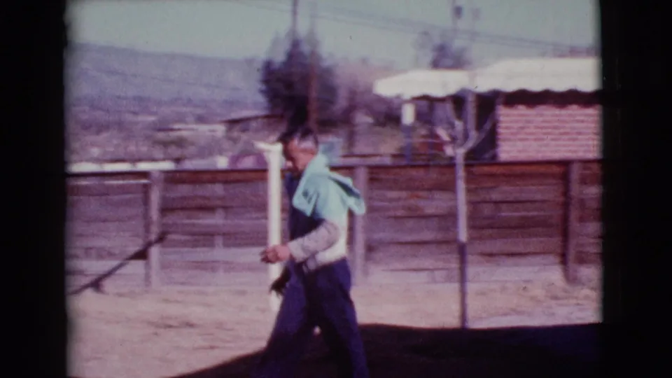 A man walks across the Cottonwood yard with a light cloth over his shoulders, passing fencing and a brick wall with hills faintly visible beyond.