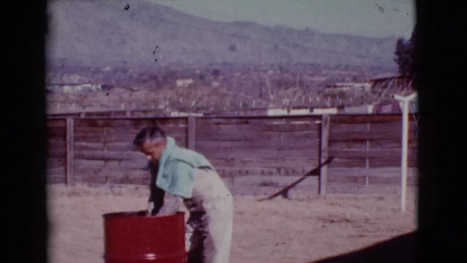 A man in light coveralls bends into a red barrel in a Cottonwood backyard, with a fence, brick wall, patio chair, and distant hills visible.