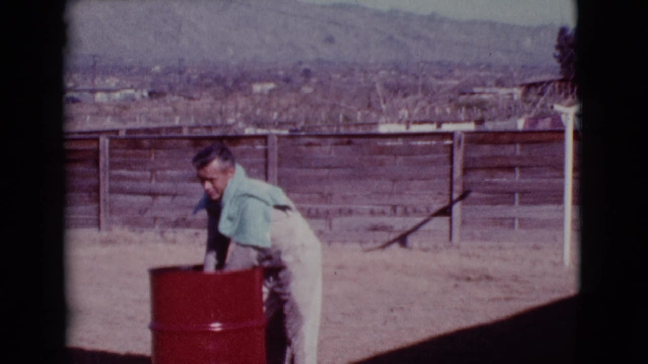 Man in light clothing stands by a red barrel in a sunny yard.