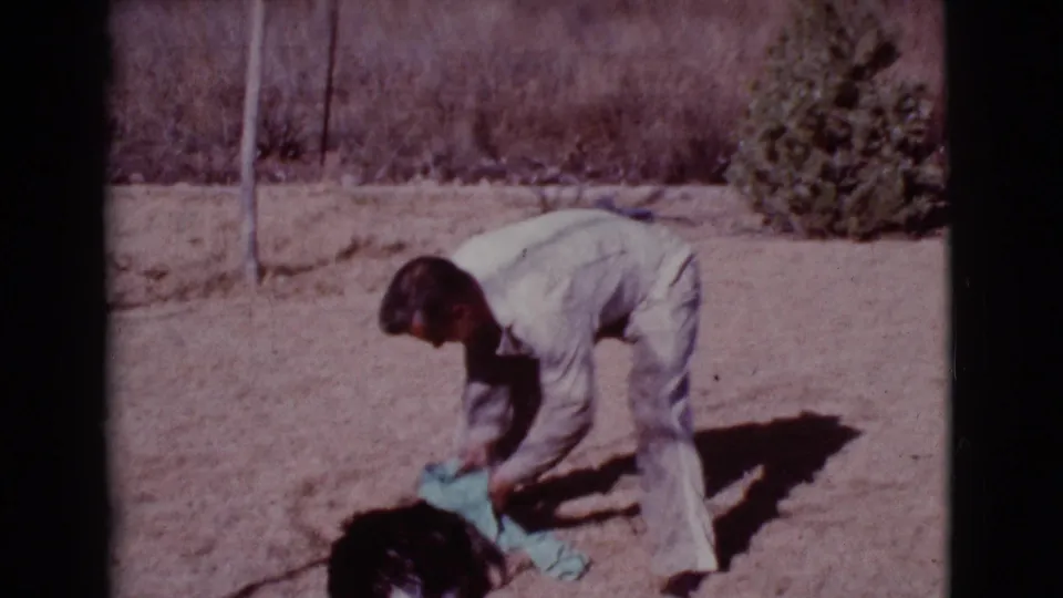 A man in light clothing bends over a black dog with a blue towel in a sunny Cottonwood, Arizona yard in 1968.