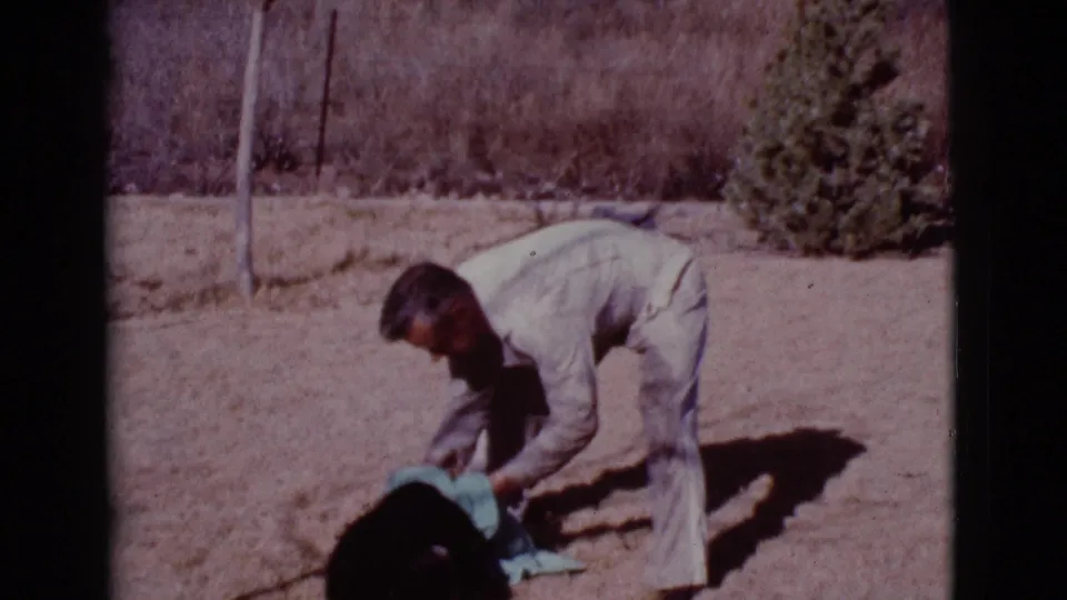 The man continues tending to the black dog with a blue towel in the dry backyard setting documented in the 1968 clip.