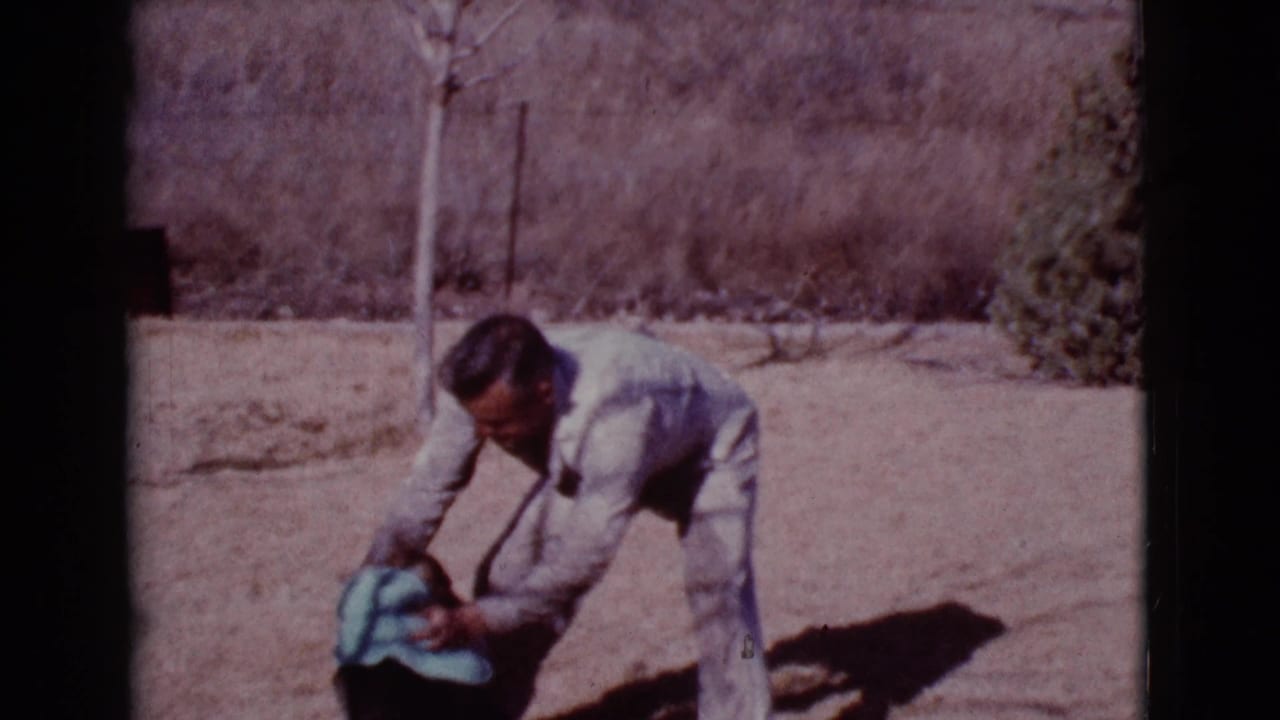 Man bends toward a dog with a blue towel in a dry sunlit yard.