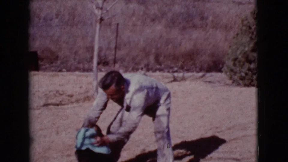 In Cottonwood, Arizona, 1968 footage, a man in light clothing bends over a German Shepherd with a blue towel in a dusty yard.