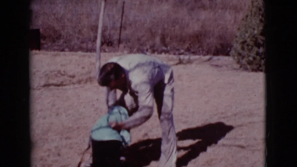A man in light work clothes towels a German Shepherd lying calmly on the ground in sunlit 1968 Cottonwood, Arizona home-movie footage.