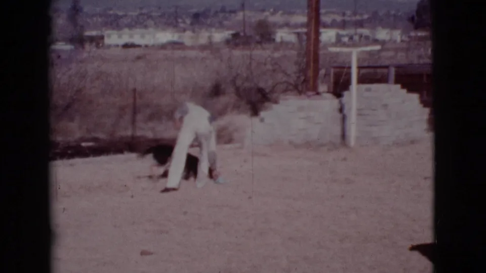 In Cottonwood, Arizona home-movie footage from 1968, a man in white bends toward a dog near dry grass and distant houses.