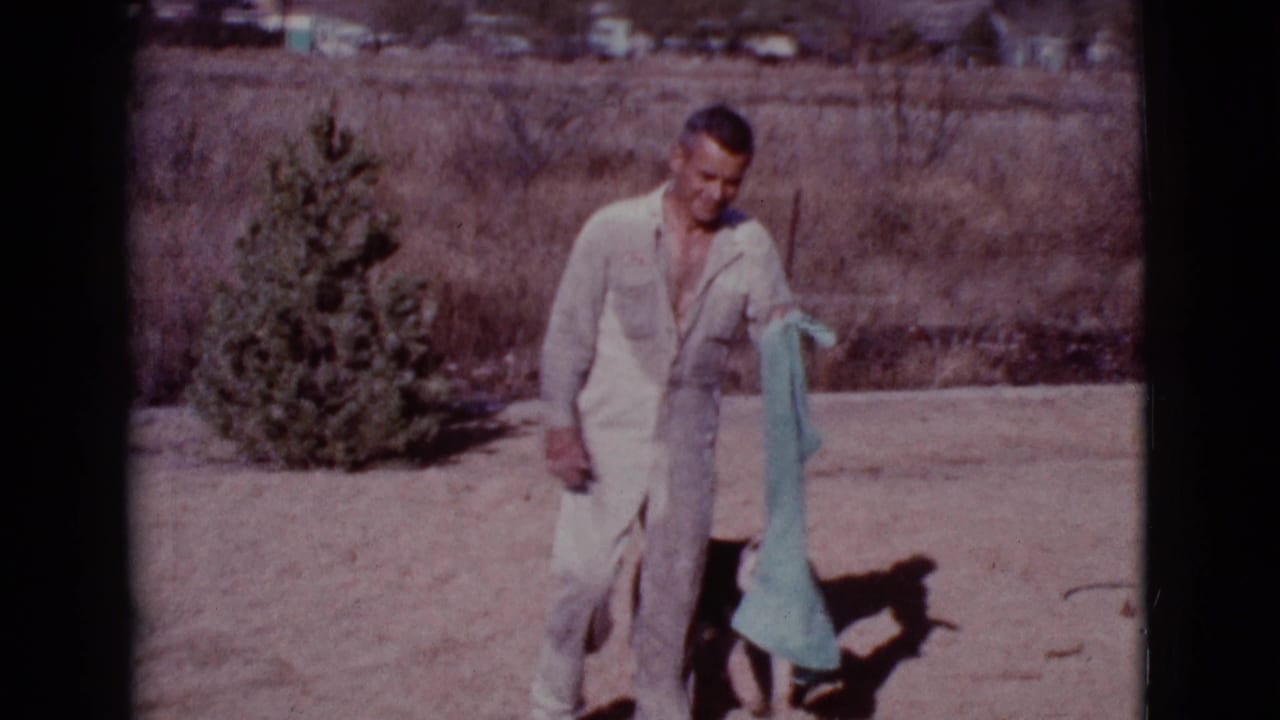 Man in a pale jumpsuit holds a teal towel in a dry yard.