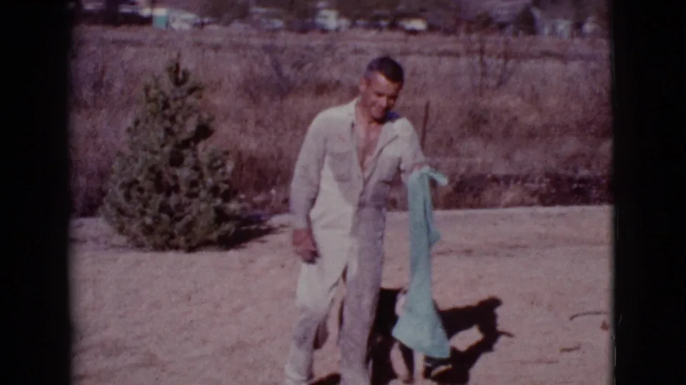 In Cottonwood, Arizona, 1968, a man in a pale jumpsuit stands outdoors holding a teal towel near dry grass and a small evergreen.