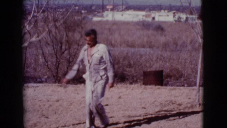 A man in light clothing crosses the dry Cottonwood, Arizona yard in this 1968 8mm frame, with sparse trees and distant buildings behind him.