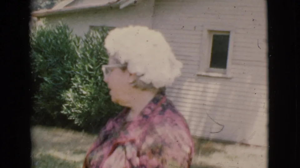 Older woman in glasses and a patterned pink jacket stands in a Cottonwood yard near shrubs and a house exterior.