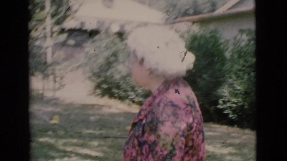 Older woman with white hair is filmed from the side in a grassy Cottonwood yard with trees and shrubs behind her.