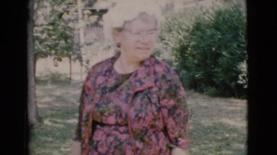 Older woman in glasses and a patterned pink jacket faces the camera in a 1968 Cottonwood residential yard scene.