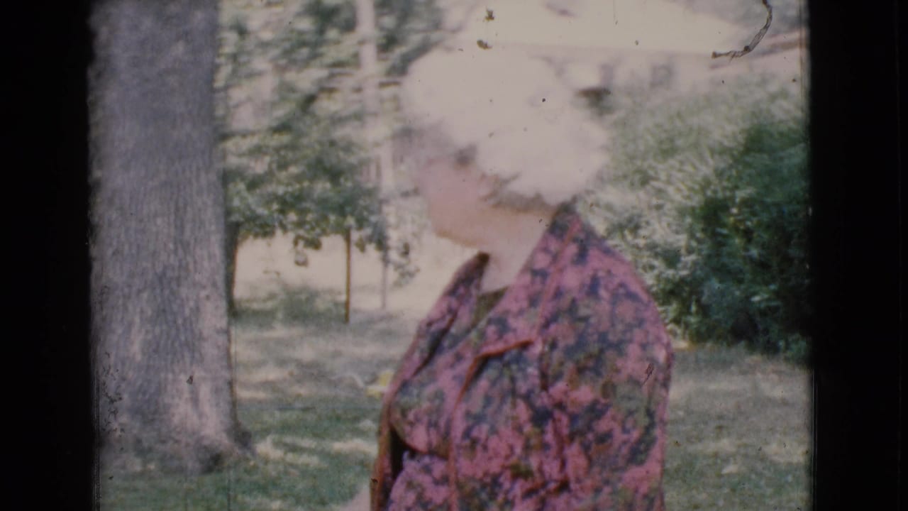 Older woman in a floral dress standing in a yard beside a tree.
