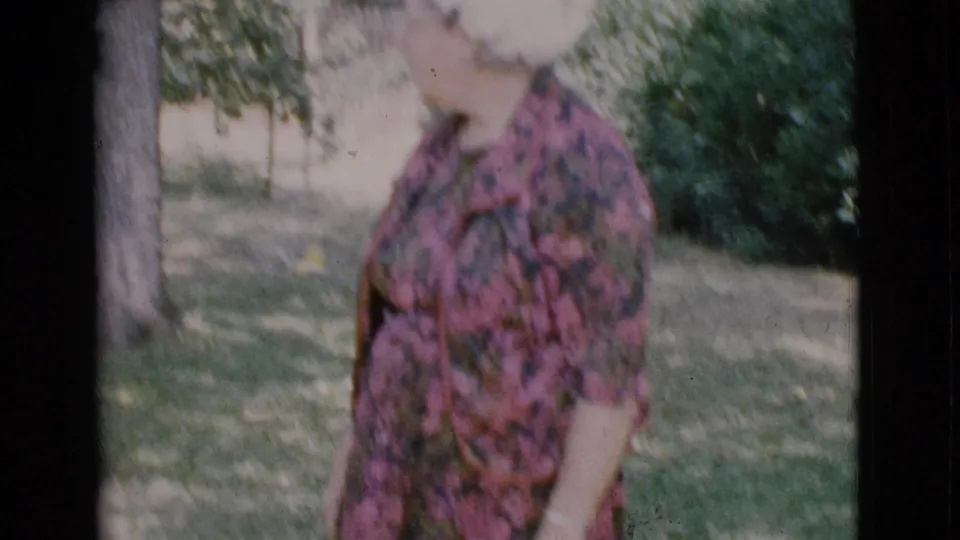 The woman stands outdoors in the Cottonwood yard, with trees, grass, and shrubs behind her in faded 8mm color.