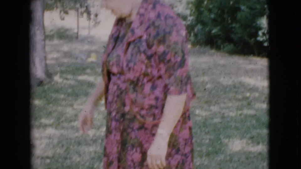In the 1968 Arizona home-movie frame, the woman in a rose floral dress bends slightly while standing on the lawn.