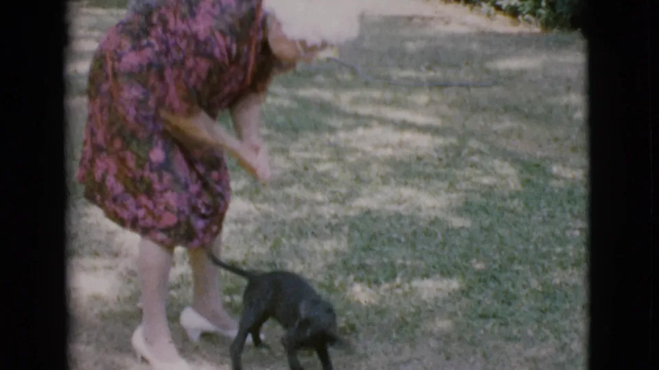 A woman in white heels leans over a small black puppy near her feet during a brief 1968 yard play moment.