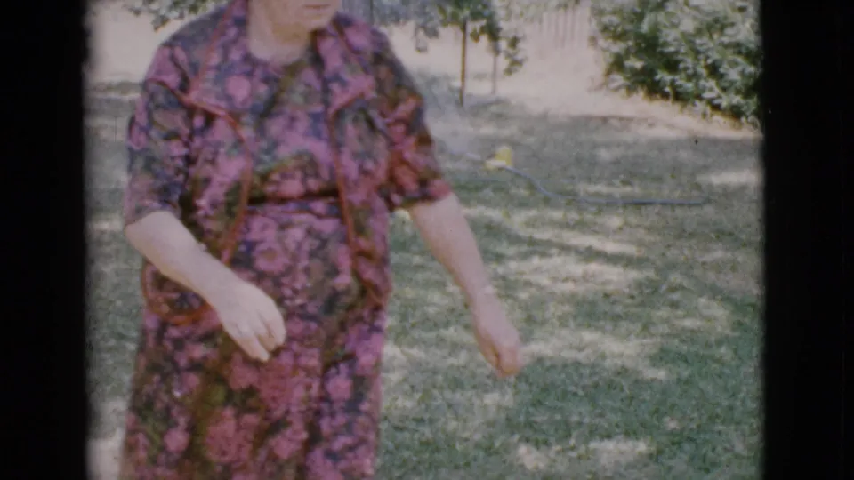 A woman in a pink and green patterned dress walks through the Cottonwood yard, with the home-movie camera cropping her face and lower body.