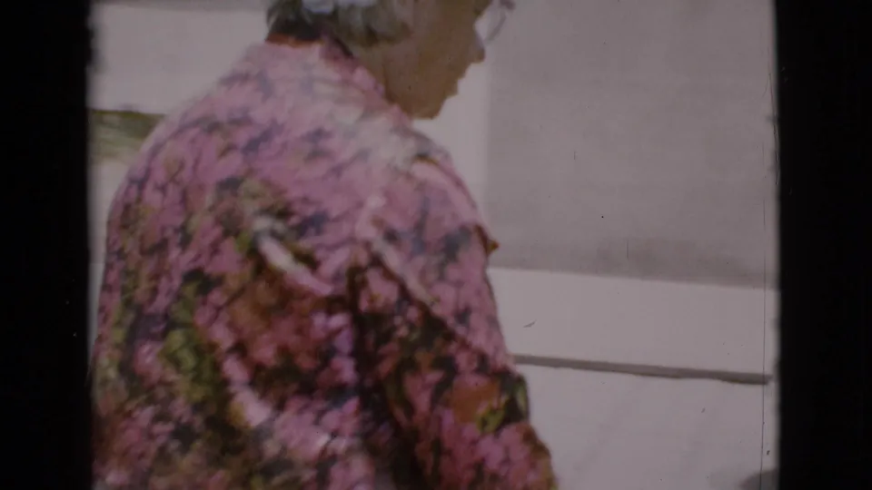 Side view of the elderly woman walking near white house siding in 1968 Cottonwood, Arizona home-movie footage.