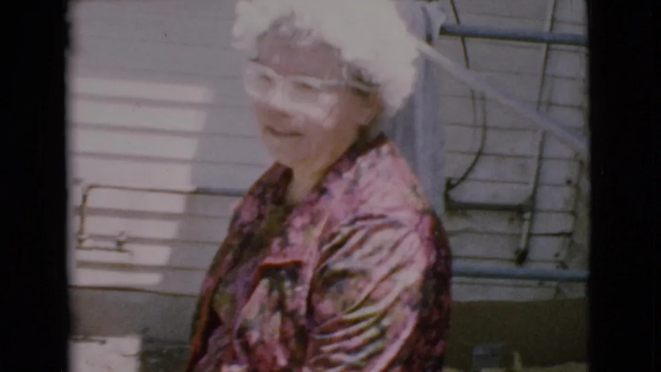 Elderly woman with glasses and white hair smiles near a house exterior in 1968 Cottonwood, Arizona home-movie footage.