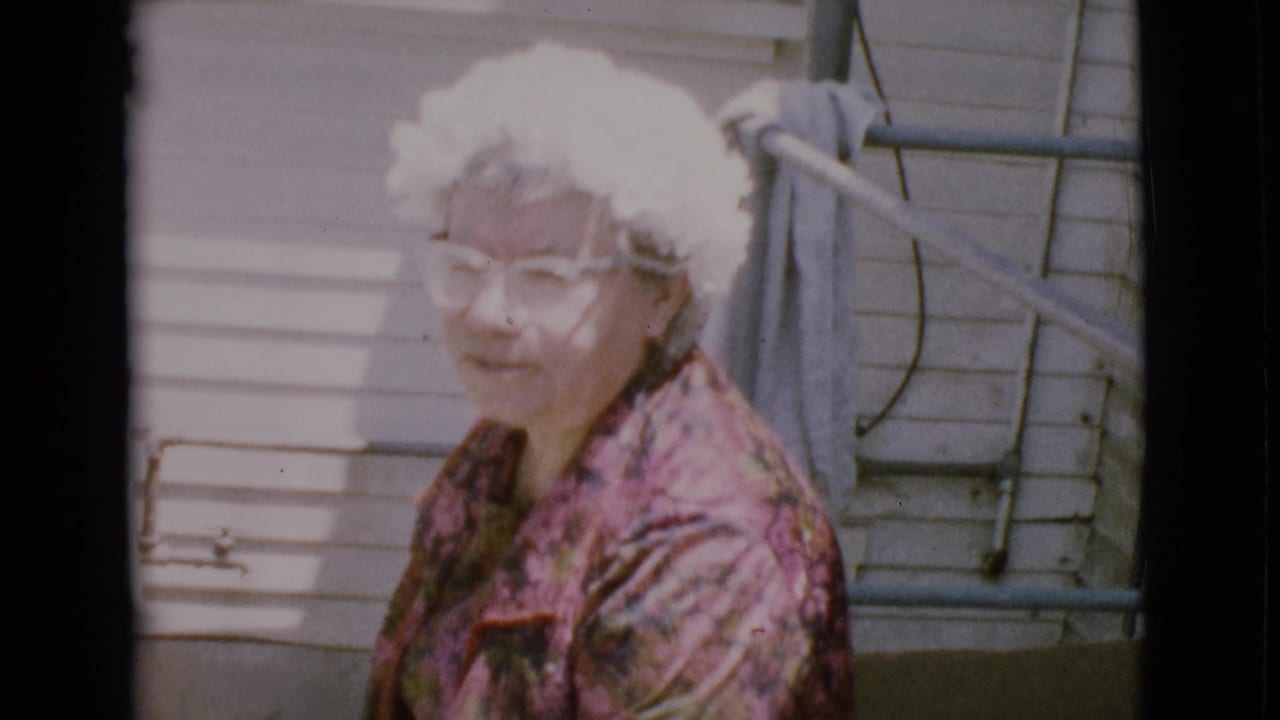 Older woman in glasses and floral dress seated or crouched outside a house.