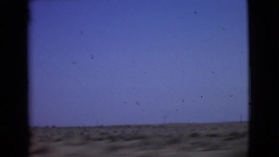 Brief 1968 San Diego County desert view, likely from a moving vehicle, with blue sky, low scrubby ground, dark film edges, and surface wear.
