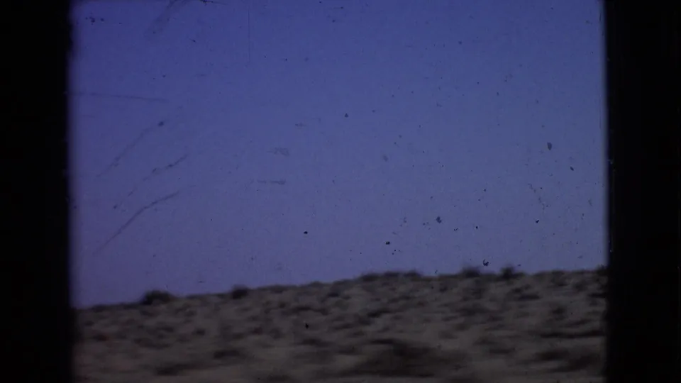 Passenger-window view of arid San Diego County terrain in 1968, with scrubby ground, blue sky, and visible 8mm film marks.